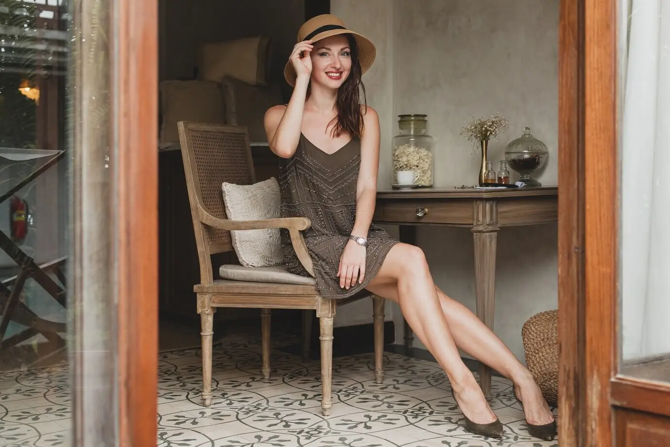 Junge, schöne, stilvolle Frau im Zimmer eines Resort-Hotels, sitzt am Tisch, trägt ein trendiges Kleid im Safari-Stil und einen Strohhut, lächelnd, glücklich, Sommerurlaub, Boho-Outfit.