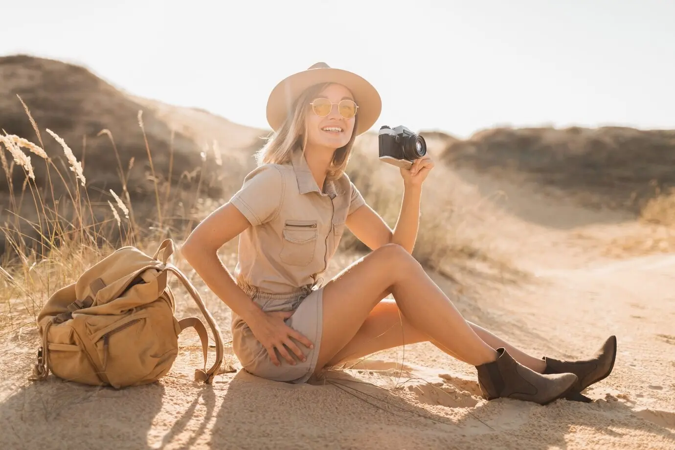 Eine attraktive, stilvolle junge Frau in einem khakifarbenen Kleid befindet sich in der Wüste, ist auf Safari in Afrika unterwegs, trägt Hut und Rucksack und macht ein Foto mit einer Vintage-Kamera.